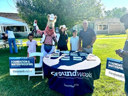 People and a mascot stand by a table with brochures at an outdoor event for Groundworks, a foundation expert company.