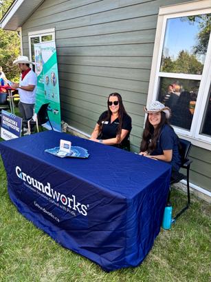 Two people sitting at a "Groundworks" table outdoors, with informational materials displayed.