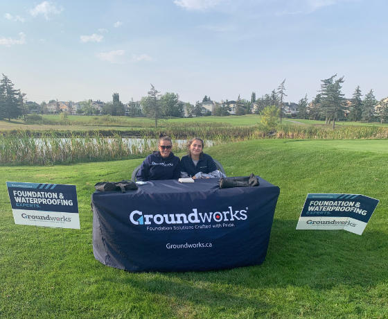 Two people sit at a table with a Groundworks tablecloth, surrounded by signs in a grassy outdoor setting, representing a Calgary charity organization dedicated to making positive change.