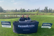 Two people sit at a table with a Groundworks tablecloth, surrounded by signs in a grassy outdoor setting, representing a Calgary charity organization dedicated to making positive change.