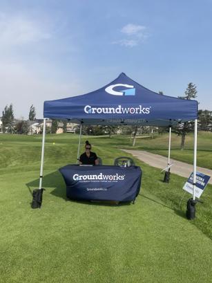A person sits at a promotional Groundworks tent on a grassy area under a clear sky, representing a Calgary charity's heartwarming efforts.