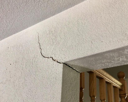 A cracked wall near a wooden staircase railing in a residential interior.