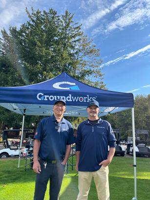 Two men stand smiling under a blue Groundworks tent at a golf course with trees and clear sky.