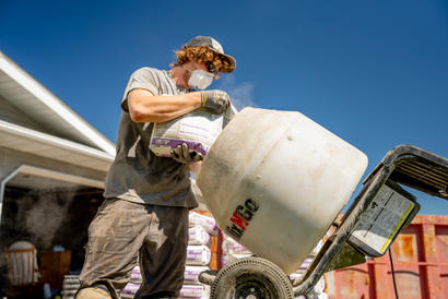 Person wearing a cap and mask pours a bag of cement mix into a rotating mixer under a clear sky.