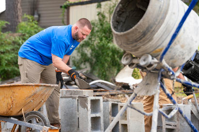 A man in a blue shirt laying mortar on cinder blocks next to a cement mixer and wheelbarrow outdoors.