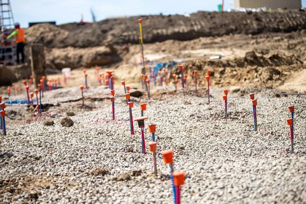 Gravel-covered construction site with many orange-topped stakes marking locations on the ground.