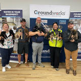 Five smiling people stand holding puppies in front of Groundworks promotional banners indoors.
