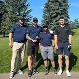 Four men stand together on a golf course in sunny weather, wearing golf attire and smiling at the camera.