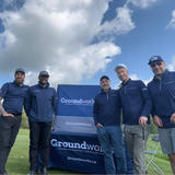 Five men in blue jackets stand smiling by a Groundworks sign outdoors on a sunny day with clouds.