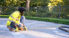 Person in a yellow safety vest measures distance on a paved path with a tape measure outdoors.