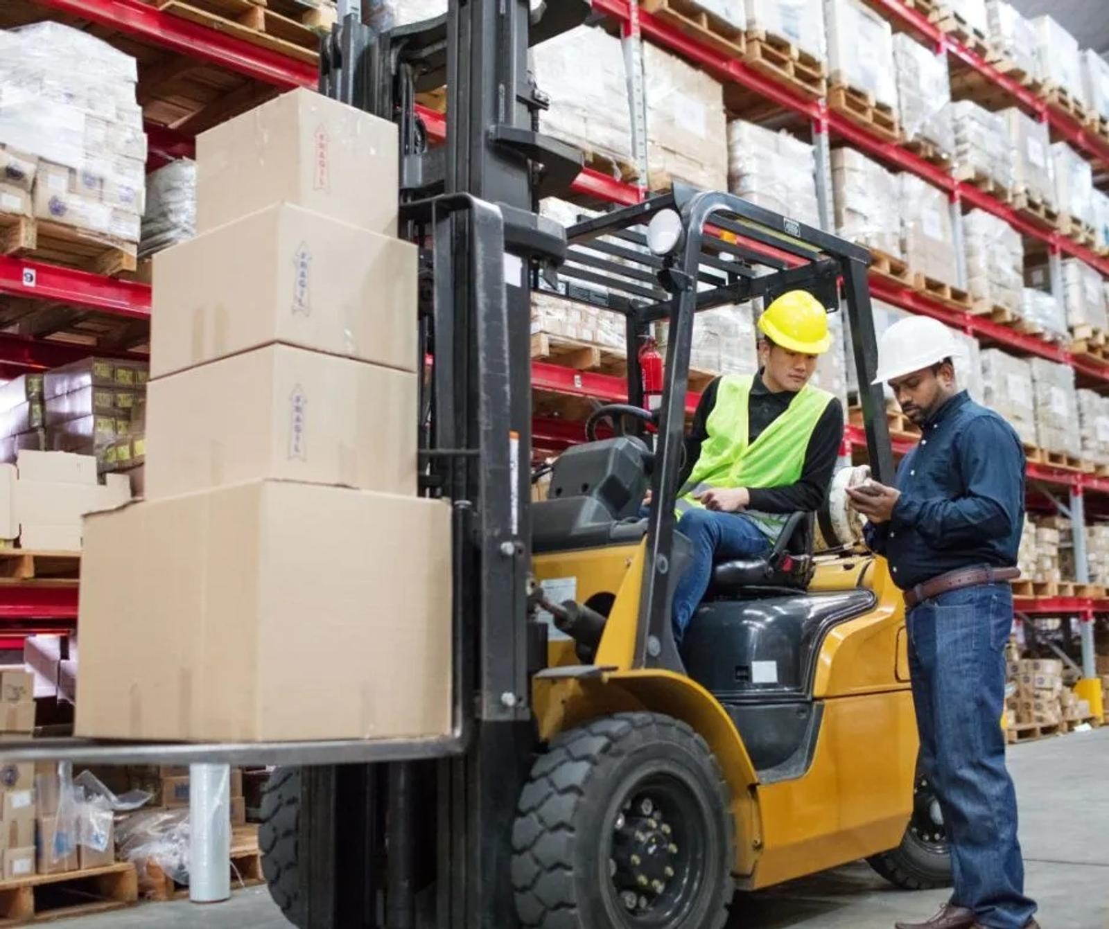 Two workers with safety gear operate a forklift in a warehouse stacked with boxes on shelves.