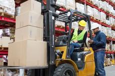 Two workers with safety gear operate a forklift in a warehouse stacked with boxes on shelves.