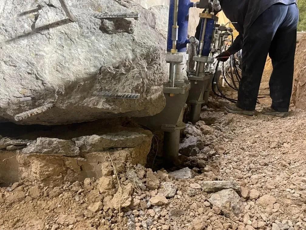 A worker operates machinery to lift a large rock slab from uneven, rocky ground.
