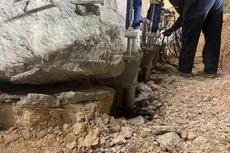 A worker operates machinery to lift a large rock slab from uneven, rocky ground.