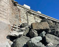 Large rocks piled at the base of a weathered concrete wall under a clear blue sky.