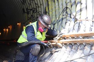 Construction worker in safety gear installs wooden slats on a tunnel wall, surrounded by tools and lights.