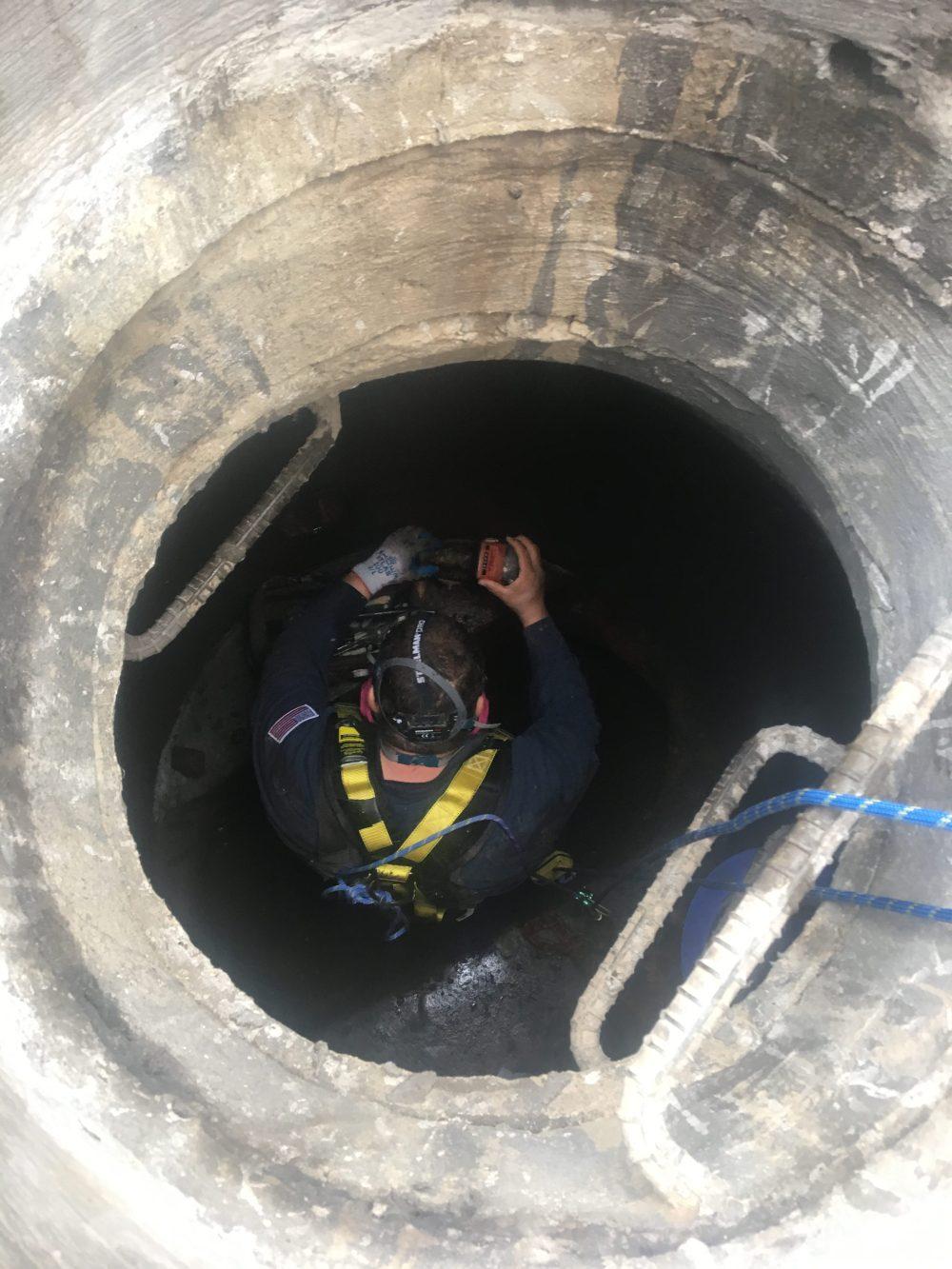 A worker in safety gear descends into a large, circular manhole using a rope and ladder.