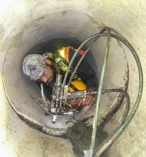 A worker in safety gear operates equipment inside a narrow concrete pipe or tunnel.