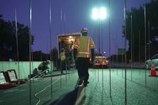 Construction workers in safety vests work on a road at night under bright lights with traffic cones nearby.