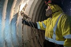 Worker in yellow safety gear uses a tool to inspect or repair the inside wall of a metal tunnel.
