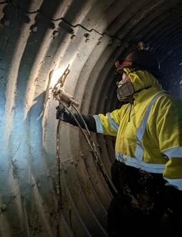 Worker in yellow safety gear uses a tool to inspect or repair the inside wall of a metal tunnel.