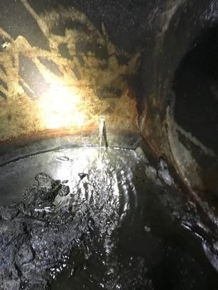 A small jet of water leaks upward from a crack in a rusty, dirty metal surface, creating ripples.