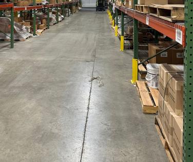 Warehouse aisle with shelves stacked with boxes and supplies on both sides, concrete floor visible.