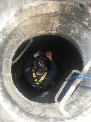 Worker wearing safety gear descends into a large, circular concrete shaft with a rope and ladder.