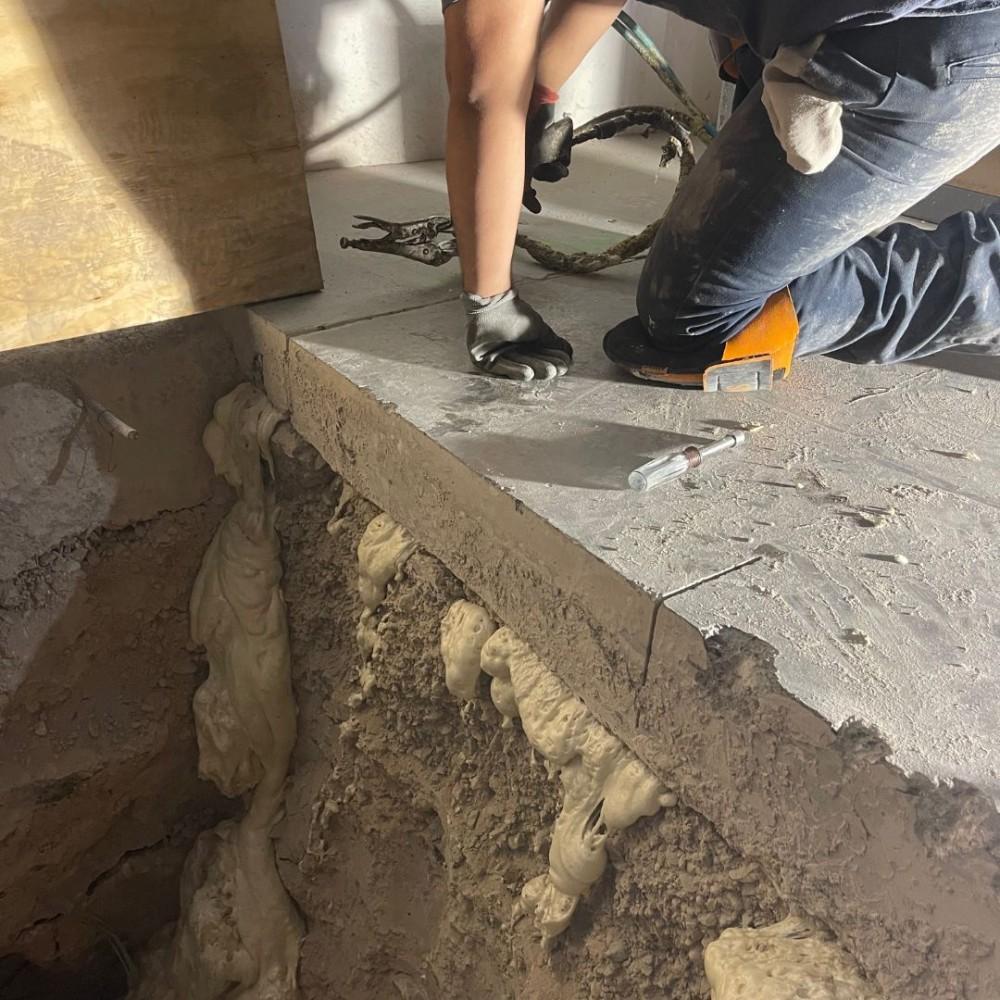 Person kneeling on concrete, working near a large crack filled with spray foam insulation below.
