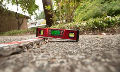 A digital level placed on cracked concrete shows the surface is uneven, with greenery in the background.
