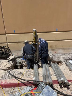 Two workers in hats and dark clothing operate machinery in a trench beside a tan building with metal pipes nearby.