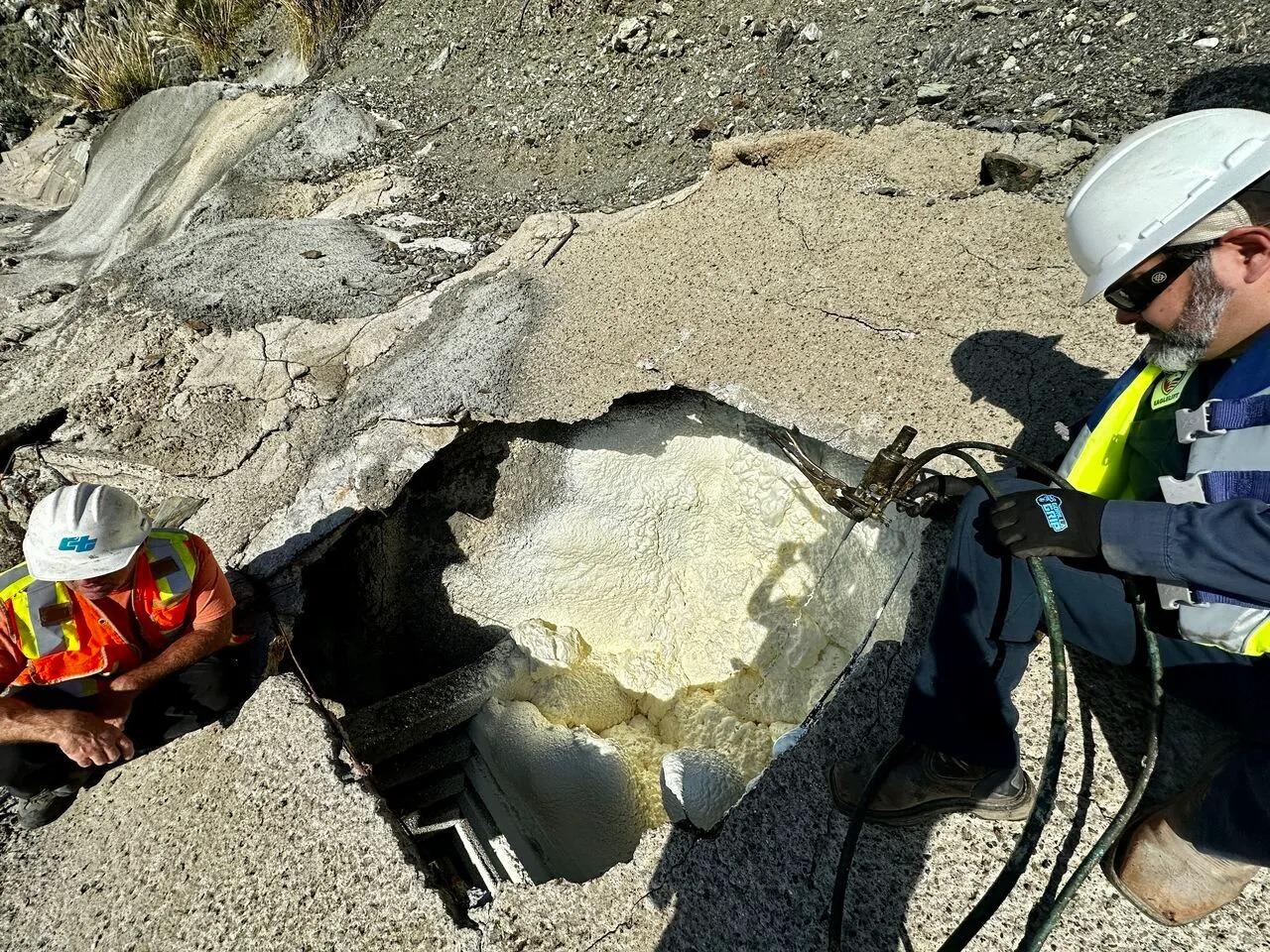 Two workers repair a large hole in concrete, injecting material while wearing safety gear and helmets.