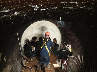 Worker in safety gear applies coating inside a large, dark tunnel using specialized equipment.