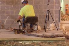 Construction worker in a yellow shirt sits on a chair near a brick wall, using equipment on the ground.