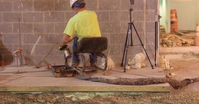 Construction worker in a yellow shirt sits on a chair near a brick wall, using equipment on the ground.