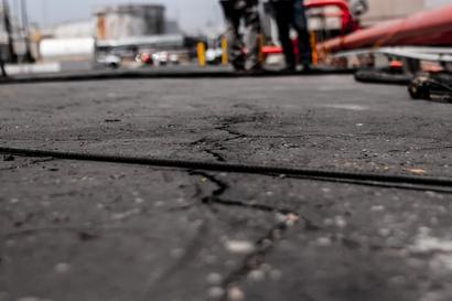 Close-up of cracked asphalt on a road, with blurred figures and industrial equipment in the background.