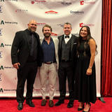 Four people dressed formally pose together on a red carpet in front of a sponsor-branded backdrop.
