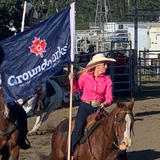 Woman in pink shirt rides horse, holding "Groundworks" flag at an outdoor rodeo event.