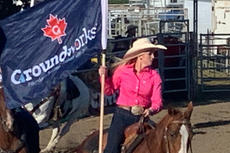 Woman in pink shirt rides horse, holding "Groundworks" flag at an outdoor rodeo event.