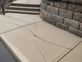 Concrete sidewalk with multiple large cracks near stairs and a stone wall.