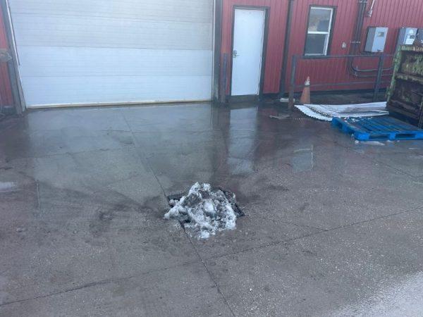 Small pile of dirty snow melting on a wet concrete surface near a red building with a garage door.