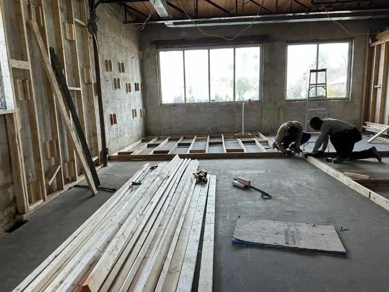 Two people working on wooden framing inside a room under construction with large windows in the background.