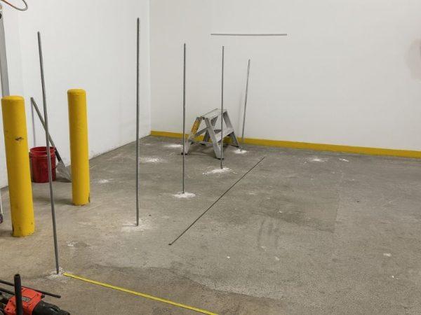 Six metal rods are installed upright in a concrete floor near a step ladder, with yellow bollards at the side.