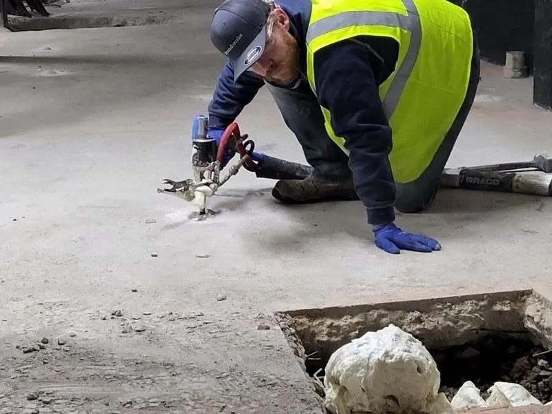 A worker in a yellow vest uses a tool near a hole in the ground with a skull inside.