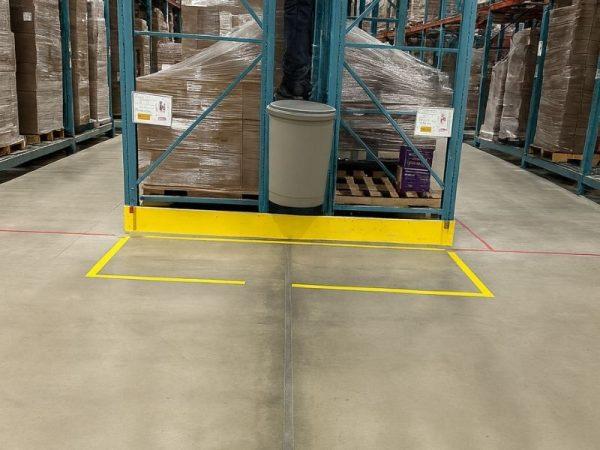 A person stands on a trash can between warehouse shelves with pallets and boxes in the background.
