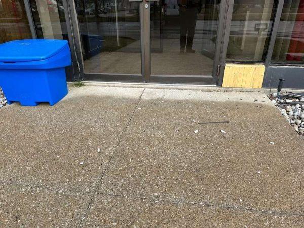 A glass door with a reflection, blue bin on left, scattered debris, and a wooden plank on the right side.