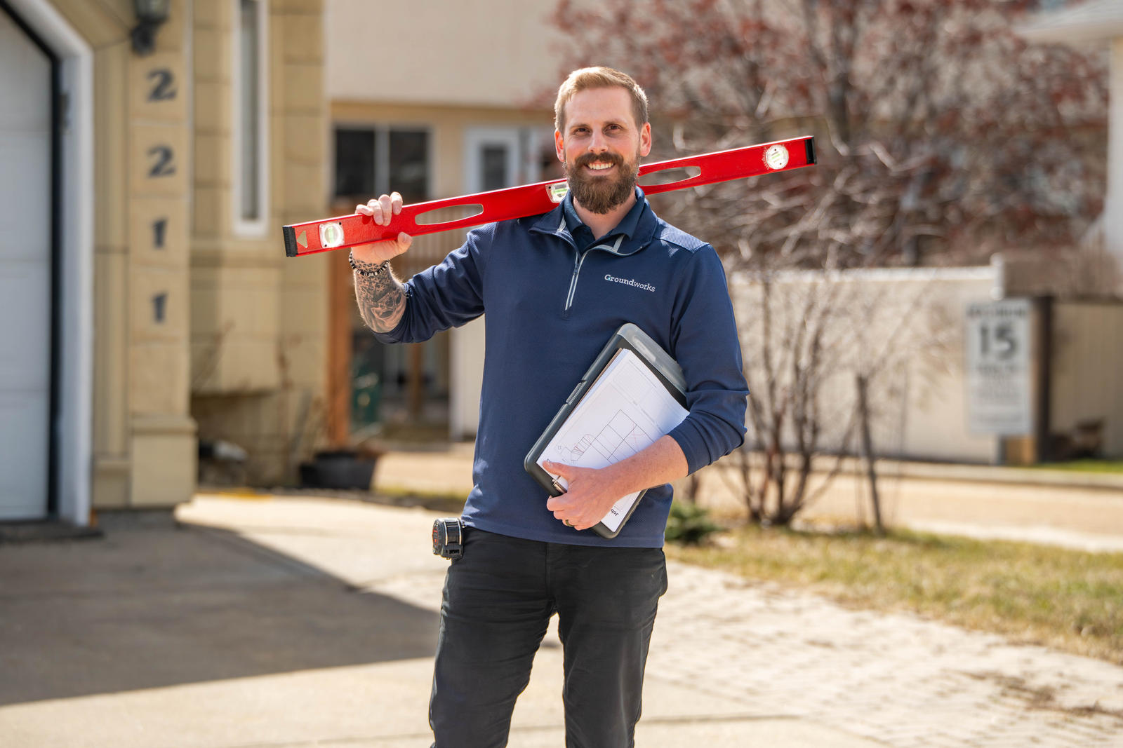 A smiling man holds a red level and blueprints, standing outside a house on a sunny day.