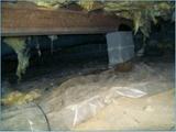 Crawl space with exposed insulation, wooden beams, and a concrete support block. Dust and plastic sheeting on the ground.