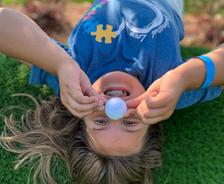 Child upside down on grass, smiling, holding a ball on their nose.