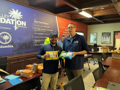 Two men smiling in an office while holding various packaged items.